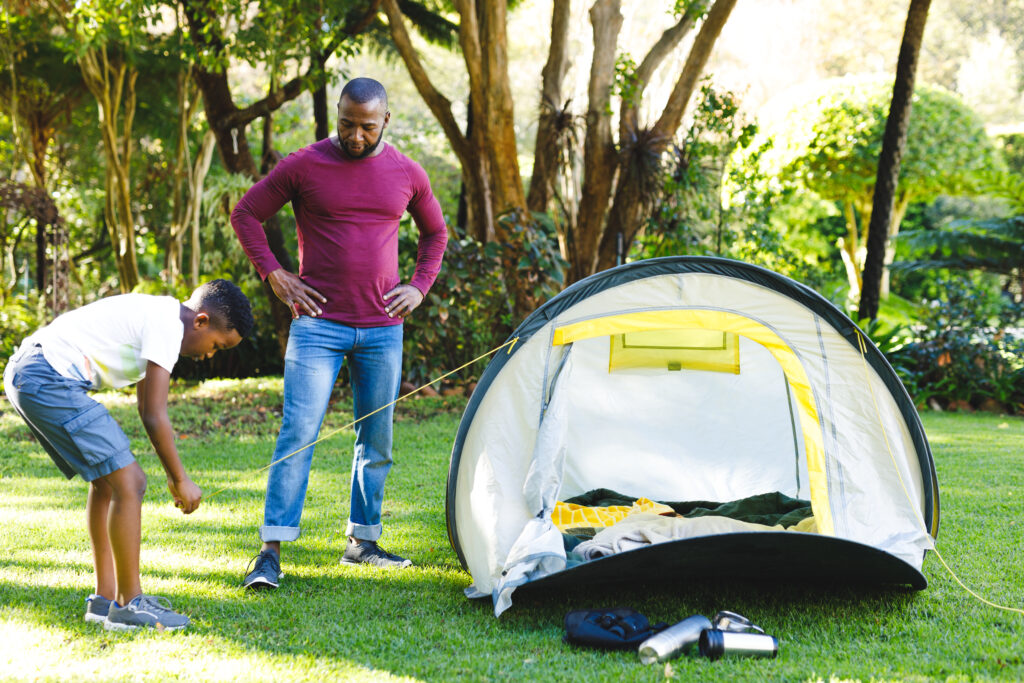 African American father watching son pitching tent in sunny garden. family spending time at home.