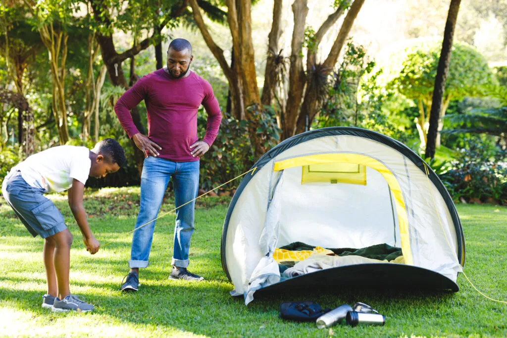 African American father watching son pitching tent in sunny garden. family spending time at home.