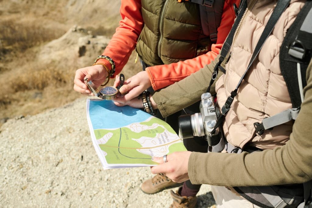 Unrecognizable female tourists wearing backpacks hiking together on sunny spring day reading paper map and using compass to find direction