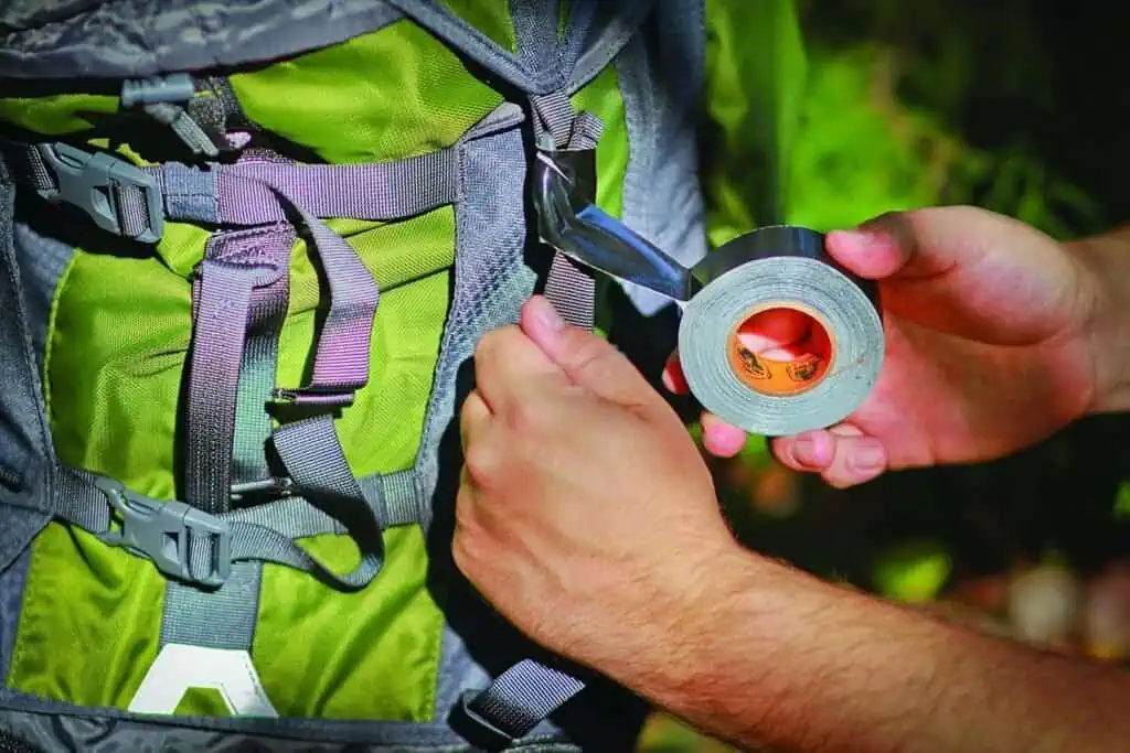 Close-up of a person attaching paracord to a backpack with duct tape, essential survival tools for preppers in outdoor emergencies.