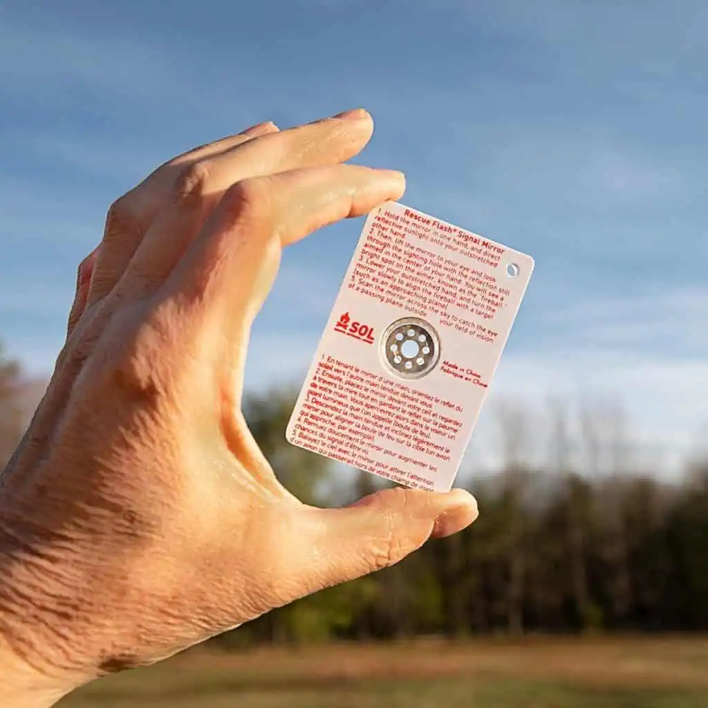Hand holding a rescue flare signal mirror outdoors with a clear sky, used for emergency signaling in survival situations.