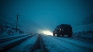 A rugged SUV driving on a snow-covered road at night, illuminated by headlights, with a winter landscape and power lines in the background, emphasizing winter vehicle safety.
