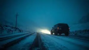 A rugged SUV driving on a snow-covered road at night, illuminated by headlights, with a winter landscape and power lines in the background, emphasizing winter vehicle safety.