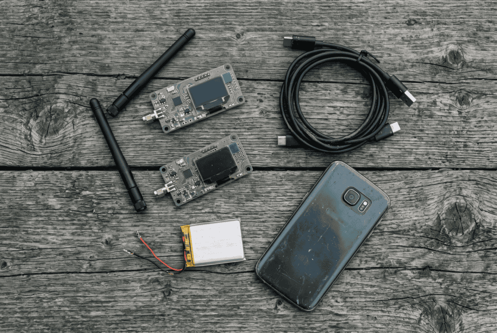 Meshtastic setup kit laid out on weathered wood — two LoRa boards, two whip antennas, a USB-C data cable and a lithium battery